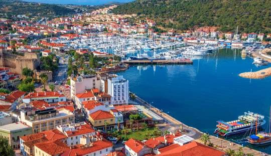 Aerial view of a Mediterranean harbor town with red-roofed buildings, blue waters filled with sailboats and yachts, surrounde